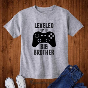 Może przedstawiać: Szara koszulka z czarnym nadrukiem kontrolera do gier wideo i tekstem "LEVELED UP TO BIG BROTHER".