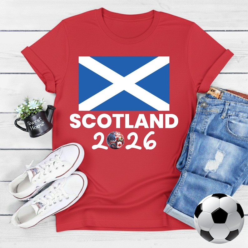 May include: Red t-shirt with the Scottish flag and the text "SCOTLAND 2026". The design includes a soccer ball with flags of the USA, Canada, and Mexico. White sneakers and a soccer ball are also in the image.