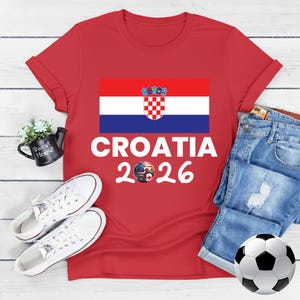 May include: Red t-shirt with the Croatian flag and the text "CROATIA 2026". The design includes a soccer ball with flags. White trainers, a football, and denim jeans are also in the image.