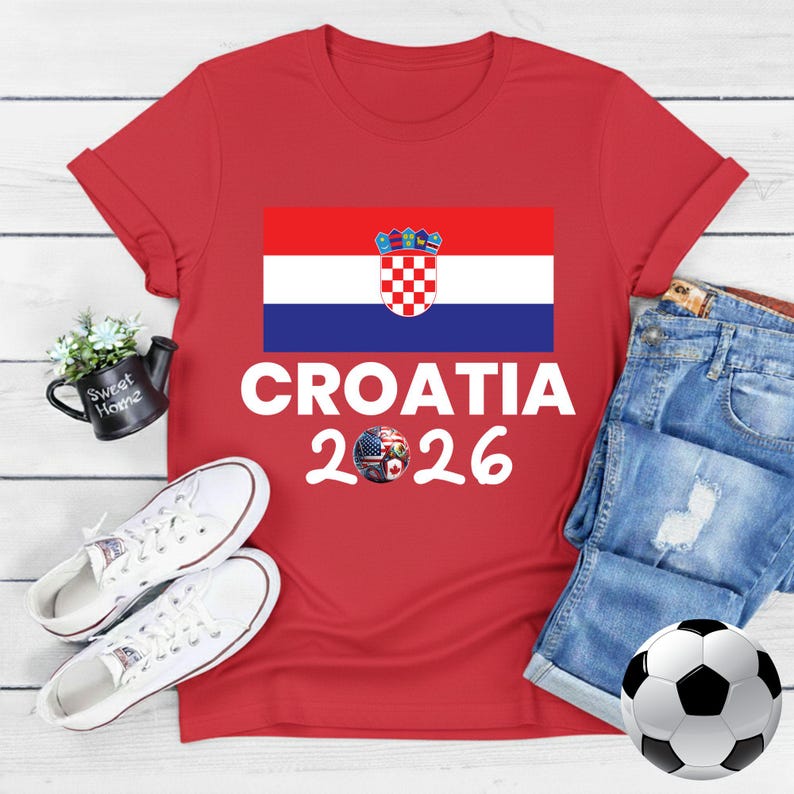 May include: Red t-shirt with the Croatian flag and the text "CROATIA 2026". The design includes a soccer ball with flags. White trainers, a football, and denim jeans are also in the image.