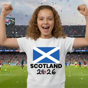 May include: A white t-shirt featuring the Scottish flag and the text "SCOTLAND 2026" in black. The shirt is worn by a person with curly hair, celebrating with arms raised. A soccer game is in the background.