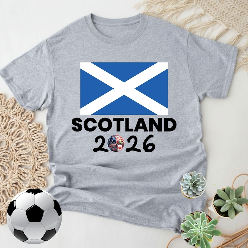 May include: A heather gray t-shirt featuring the Scottish flag and the text "SCOTLAND 2026". The design includes a soccer ball and a globe with the flags of the USA, Canada, and other countries. The shirt is a casual wear item.