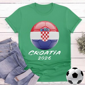 K&ouml;nnte beinhalten: Gr&uuml;nes T-Shirt mit einem Fu&szlig;ball-Grafikdesign der kroatischen Flagge und dem Text "CROATIA 2026". Eine blaue Jeans ist neben dem T-Shirt gefaltet. Ein Fu&szlig;ball befindet sich unten rechts. Ein Sportbekleidungsartikel.