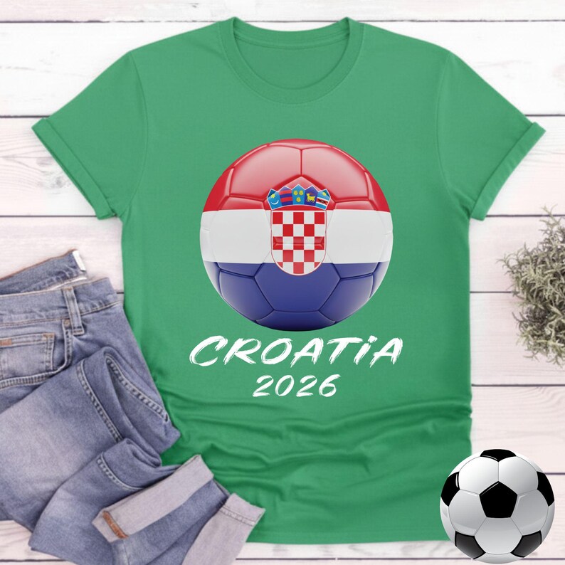 K&ouml;nnte beinhalten: Gr&uuml;nes T-Shirt mit einem Fu&szlig;ball-Grafikdesign der kroatischen Flagge und dem Text "CROATIA 2026". Eine blaue Jeans ist neben dem T-Shirt gefaltet. Ein Fu&szlig;ball befindet sich unten rechts. Ein Sportbekleidungsartikel.