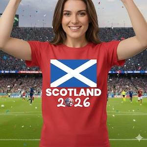 May include: Red t-shirt featuring the Scottish flag with a white cross on a blue background. Below the flag, the word "SCOTLAND" is printed in white, along with the year "2026" and a soccer ball graphic.