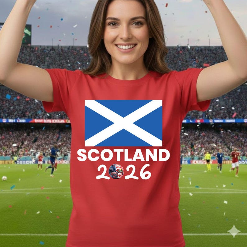 May include: Red t-shirt featuring the Scottish flag with a white cross on a blue background. Below the flag, the word "SCOTLAND" is printed in white, along with the year "2026" and a soccer ball graphic.