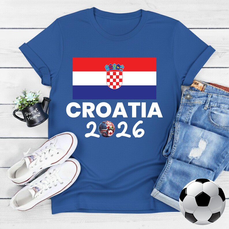 May include: Blue t-shirt featuring the Croatian flag and the text "CROATIA 2026". The design includes a soccer ball with flags. White trainers and a football are also in the image.