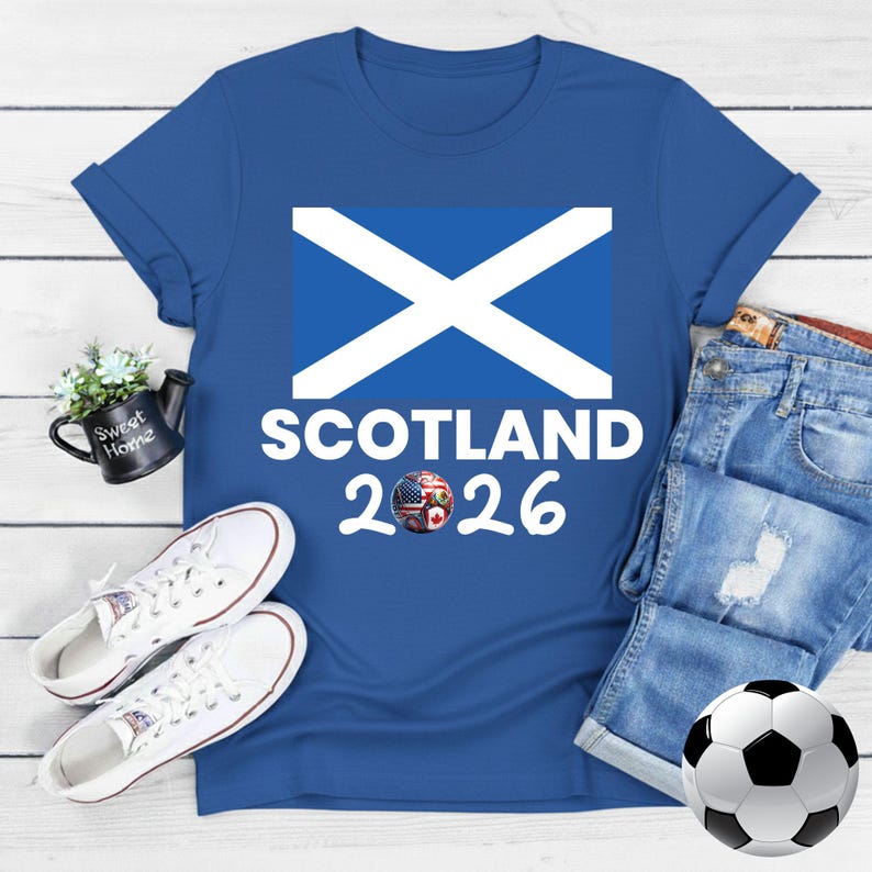 May include: A royal blue t-shirt featuring the Scottish flag and the text "SCOTLAND 2026" in white. A soccer ball with black and white panels is in the lower right corner. White sneakers and denim shorts are also in the image.
