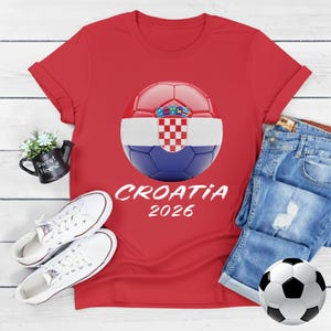 K&ouml;nnte beinhalten: Rotes T-Shirt mit einem Fu&szlig;ball-Design der kroatischen Flagge und dem Text "CROATIA 2026". Das Bild zeigt wei&szlig;e Turnschuhe und Jeansshorts. Ein Fu&szlig;ball ist ebenfalls sichtbar.