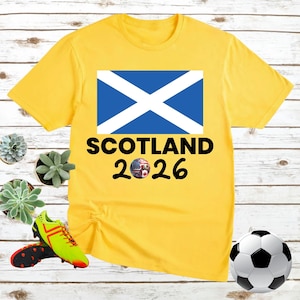 May include: A yellow t-shirt featuring the Scottish flag and the text "SCOTLAND 2026". A soccer ball and soccer cleats are also visible. The shirt has a crew neck and short sleeves.