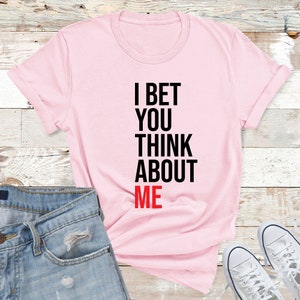 May include: Pink t-shirt with the text "I bet you think about me" in black and red lettering.
