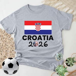 May include: A grey t-shirt with the Croatian flag and the words "CROATIA 2026". The design includes a soccer ball with the American and Croatian flags. A soccer ball and small potted plants are in the image.