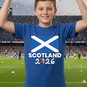May include: A blue t-shirt featuring the Scottish flag with a white X on a blue background. Below the flag, the word "SCOTLAND" is printed in white, with "2026" and a soccer ball graphic. The shirt is worn by a person in a stadium.