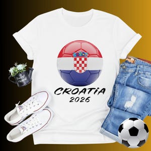 K&ouml;nnte beinhalten: Wei&szlig;es T-Shirt mit einem Fu&szlig;ball-Design mit der kroatischen Flagge und dem Text "CROATIA 2026". Das Bild zeigt auch wei&szlig;e Turnschuhe, Jeansshorts und einen Fu&szlig;ball.
