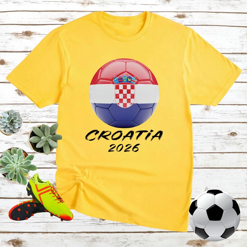 K&ouml;nnte beinhalten: Gelbes T-Shirt mit einem Fu&szlig;ball-Design der kroatischen Flagge und dem Text "CROATIA 2026". Der Ball ist rot, wei&szlig; und blau. Ein Fu&szlig;ball und Fu&szlig;ballschuhe sind ebenfalls im Bild.