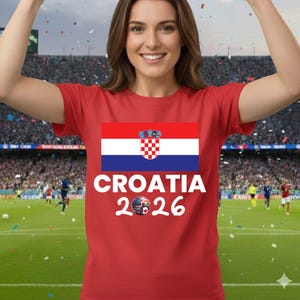May include: A red t-shirt displaying the Croatian flag and the words "CROATIA 2026" in white, with a football graphic. The shirt is worn in a stadium setting.