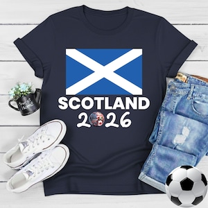 May include: Navy blue t-shirt featuring the Scottish flag and the text "SCOTLAND 2026". A soccer ball with flags of different countries is in the design. White sneakers and a soccer ball are also in the image.