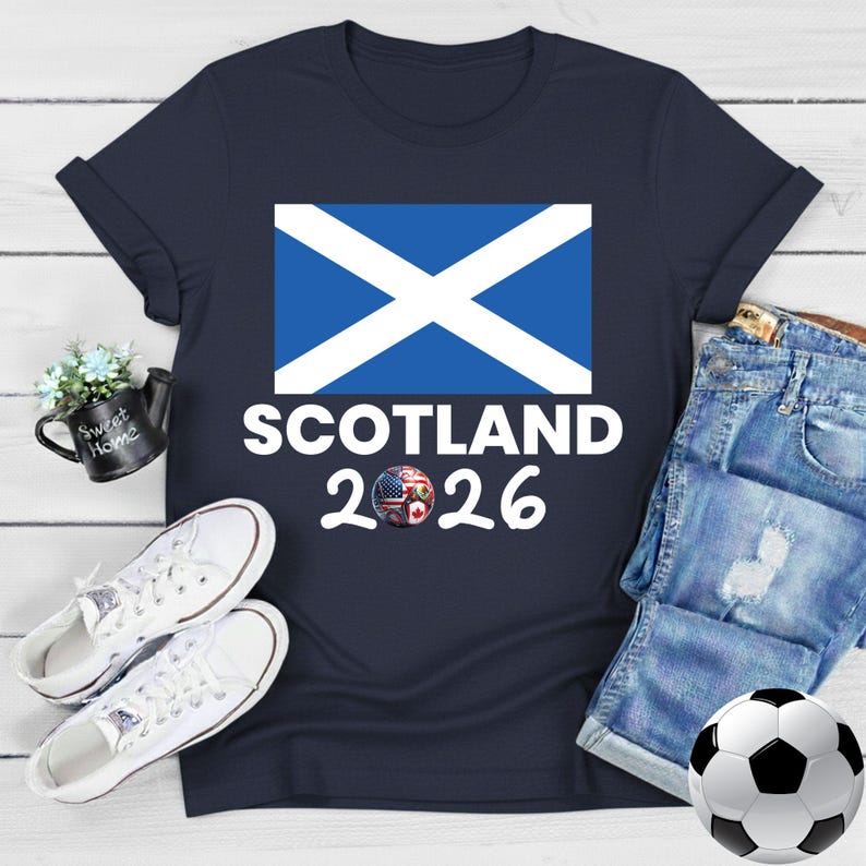 May include: Navy blue t-shirt featuring the Scottish flag and the text "SCOTLAND 2026". A soccer ball with flags of different countries is in the design. White sneakers and a soccer ball are also in the image.