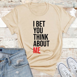 May include: A beige t-shirt with the text "I bet you think about me" printed in black and red.