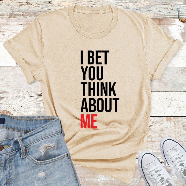 May include: A beige t-shirt with the text "I bet you think about me" printed in black and red.