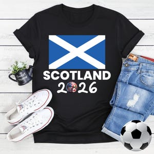 May include: Black t-shirt featuring the Scottish flag and the text "SCOTLAND 2026". The design includes a soccer ball and a graphic with the flags of the USA, Canada, and Mexico. White sneakers and denim shorts are also visible.