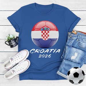 K&ouml;nnte beinhalten: Blaues T-Shirt mit einem Fu&szlig;ball im kroatischen Flaggen-Design und dem Text "CROATIA 2026". Das Bild zeigt auch wei&szlig;e Turnschuhe und einen Fu&szlig;ball. Das T-Shirt liegt auf einer Holzoberfl&auml;che.