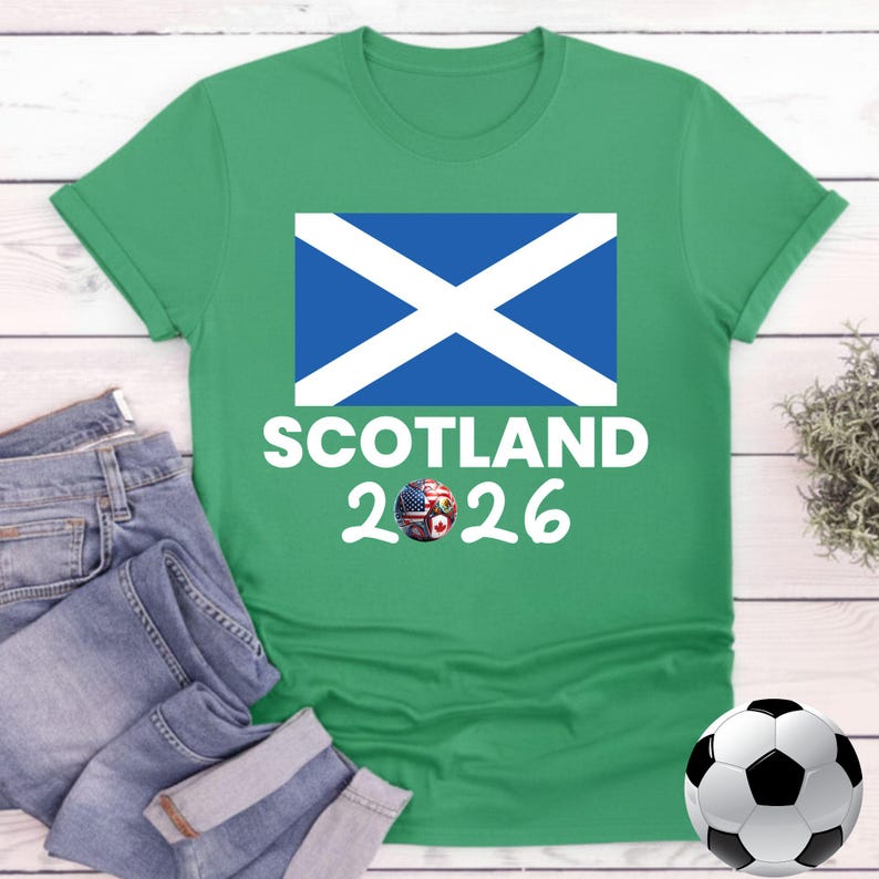 May include: A green t-shirt featuring the Scottish flag and the word "SCOTLAND" in white. Below the text is the number "2026" with a graphic of a soccer ball with the flags of the USA, Canada, and other countries. A soccer ball is in the bottom right.