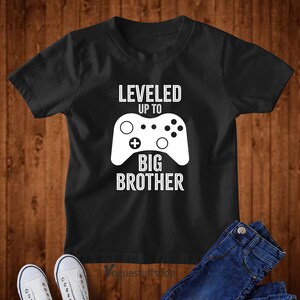 Może przedstawiać: Czarny T-shirt z białym nadrukiem kontrolera do gier wideo i tekstem "LEVELED UP TO BIG BROTHER".