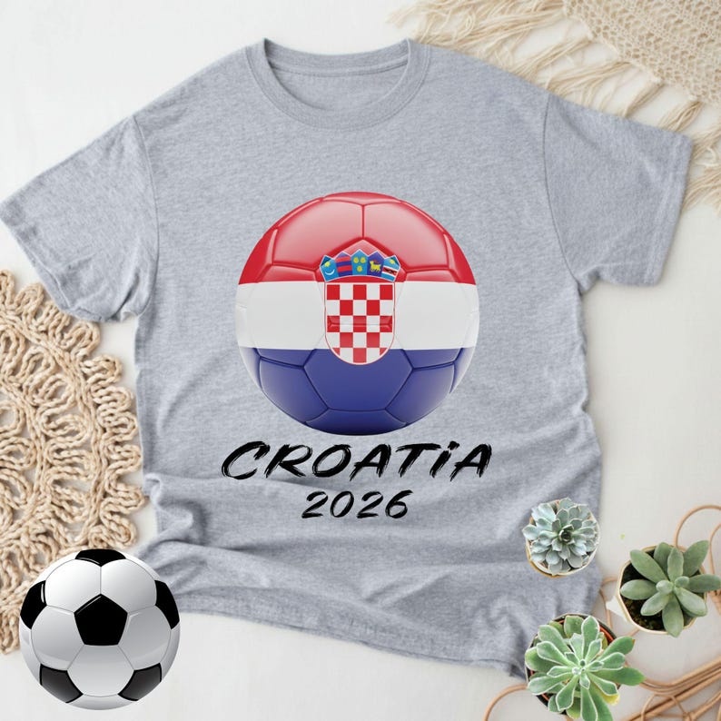 K&ouml;nnte beinhalten: Graues T-Shirt mit einem Fu&szlig;ball-Design in den Farben der kroatischen Flagge und dem Emblem. Der Text "CROATIA 2026" ist unter dem Ball aufgedruckt. Ein Fu&szlig;ball befindet sich in der linken unteren Ecke.