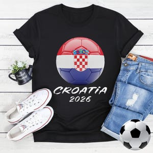 K&ouml;nnte beinhalten: Schwarzes T-Shirt mit einem Fu&szlig;ball-Design der kroatischen Flagge und dem Text "CROATIA 2026". Das Bild zeigt wei&szlig;e Turnschuhe und Jeansshorts. Ein Fu&szlig;ball ist ebenfalls vorhanden.