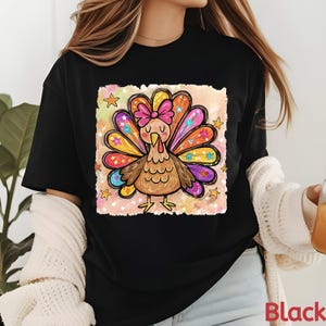 May include: A black short-sleeved t-shirt featuring a colourful graphic print of a turkey with a pink bow on its head. The turkey has vibrant, multi-coloured feathers decorated with small stars and floral patterns, set against a soft, watercolour-style background.