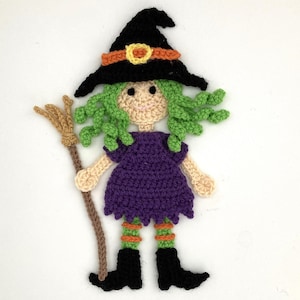 Puede incluir: Una muñeca de bruja de crochet con cabello verde, un vestido morado, medias a rayas naranja y verde y zapatos negros. La muñeca sostiene una escoba marrón.