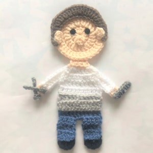 May include: A crocheted figure of a person with brown hair, a white shirt, gray pants, and blue shoes. The figure is holding a small crocheted object in each hand.