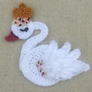May include: White crocheted swan with a crown of orange yarn and pink flowers. The swan has a red beak and pink flowers on its wings.