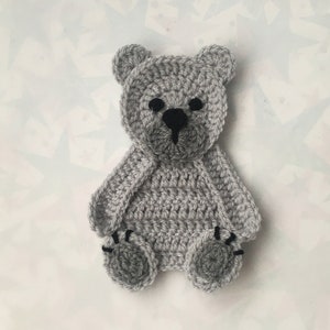 Op de afbeelding: Een grijze gehaakte teddybeer met zwarte ogen en een zwarte neus. De beer zit op een witte achtergrond met een stervormig patroon.