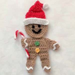 May include: A crocheted gingerbread man wearing a red and white Santa hat and holding a candy cane. The gingerbread man has a smiling face and is decorated with green and yellow buttons.