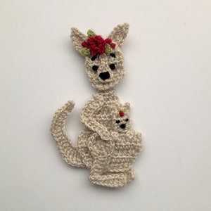 Può includere: Un canguro all'uncinetto con un piccolo nella sua tasca. Il canguro indossa una corona di fiori rossi.