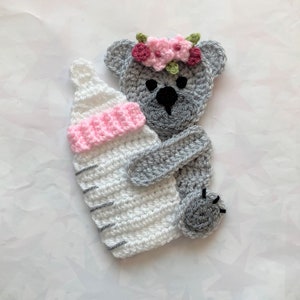 Puede incluir: Un oso de peluche gris tejido a crochet con una corona de flores rosa y verde sostiene un biberón tejido a crochet blanco y rosa.
