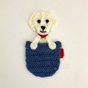 May include: A crocheted white dog with a red collar and a gold tag is peeking out of a blue crocheted pocket.