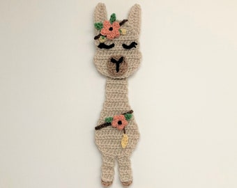 May include: A crocheted llama bookmark with a pink flower crown and a pink flower on its side. The llama is beige with brown eyes and a brown nose.
