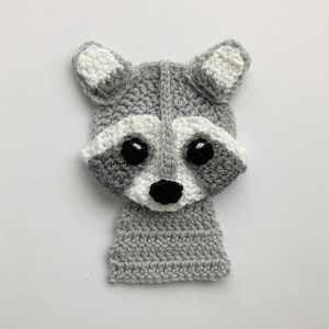 May include: A crocheted raccoon face made with grey and white yarn. The raccoon has black eyes and a black nose.