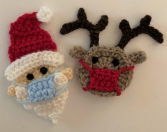 Puede incluir: Adornos de crochet de Papá Noel y reno con mascarillas faciales azules y rojas. Los adornos son blancos, rojos, marrones y azules.