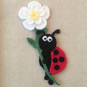 Può includere: Una coccinella all'uncinetto con dettagli neri e rossi che tiene un fiore bianco all'uncinetto con un centro giallo.