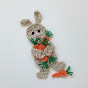 May include: A crocheted bunny holding a bunch of orange carrots with green tops. The bunny is beige and has black eyes and a pink nose.