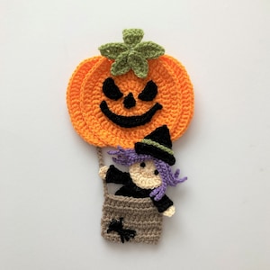 May include: A crocheted Halloween decoration featuring a witch in a purple hat and black dress riding in a hot air balloon shaped like a jack-o'-lantern.
