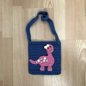 Op de afbeelding: Een blauwe gehaakte tas met een roze dinosaurus applicatie. De dinosaurus heeft gele en witte vlekken.