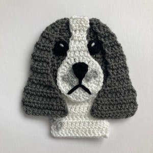 Puede incluir: Cabeza de perro de crochet en hilo gris y blanco. El perro tiene ojos negros y una nariz negra.