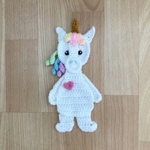 Può includere: Un unicorno bianco all'uncinetto con un cuore rosa sul petto e una criniera arcobaleno. L'unicorno è in piedi su una superficie di legno.