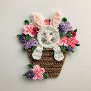May include: A crocheted white bunny rabbit peeking out of a brown flower pot with pink, purple, and green flowers.