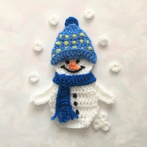 Puede incluir: Un muñeco de nieve de crochet que lleva un gorro azul y verde y una bufanda azul. El muñeco de nieve está rodeado de bolas de nieve blancas de crochet.